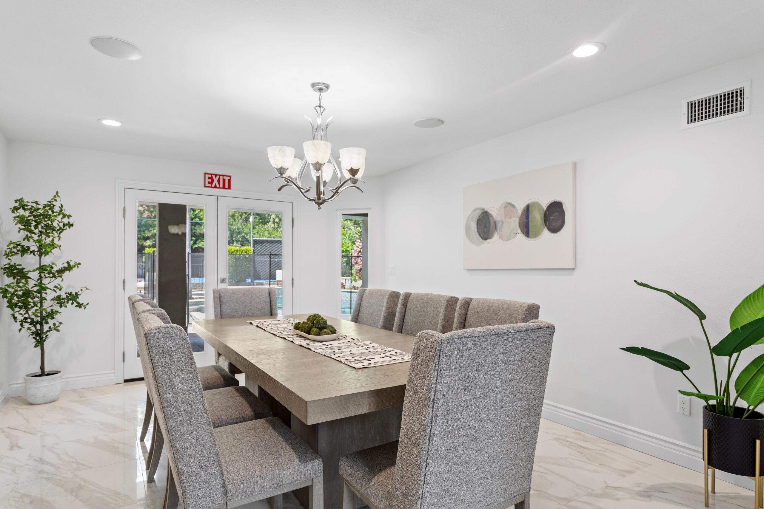 Modern dining room with a large wooden table, cushioned beige chairs, a chandelier, potted plants, wall art, tiled floor, and glass doors leading outside. The room is bright with natural light.