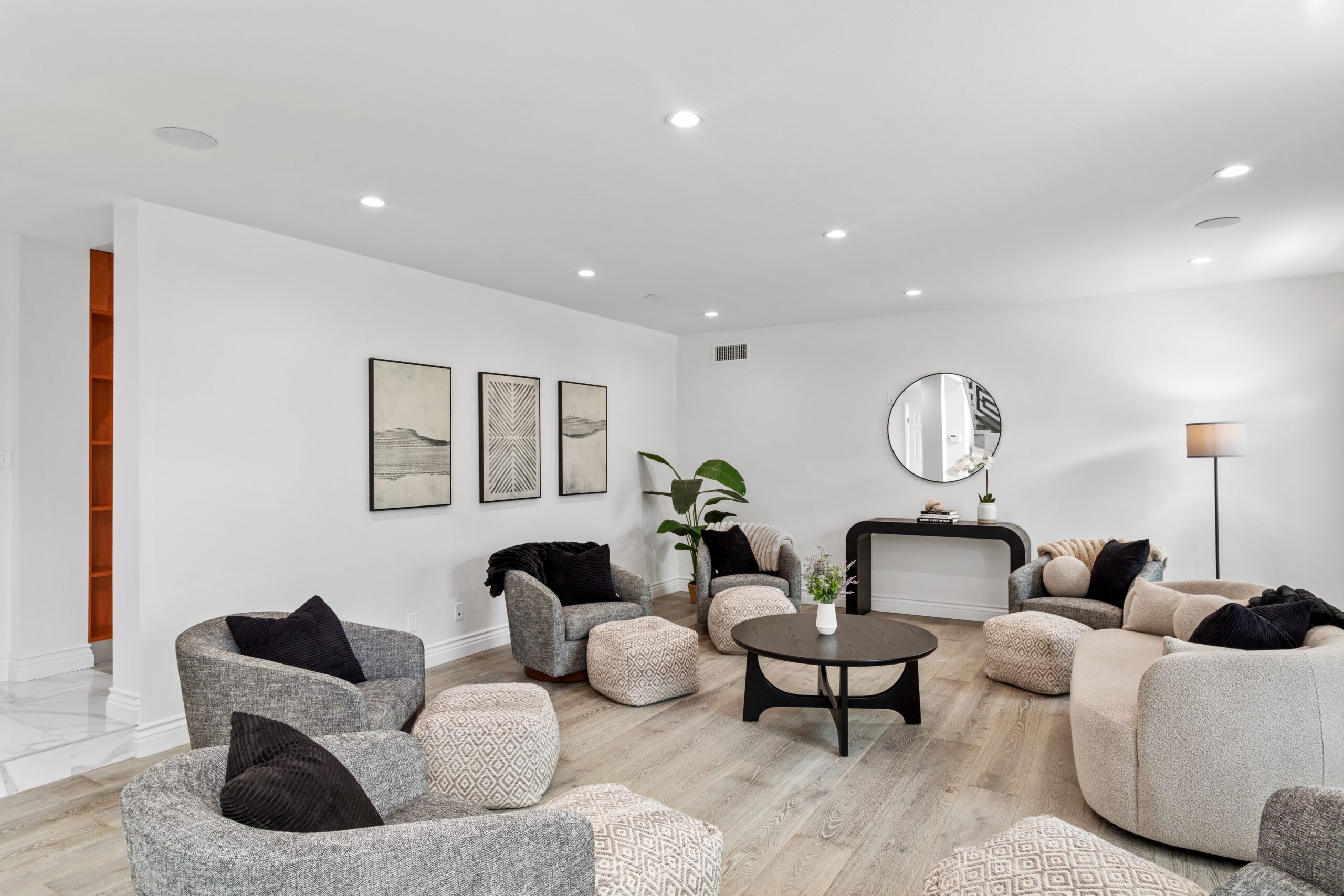 Modern living room with light wood flooring, circular gray chairs, patterned cushions, round black coffee table, wall art, a potted plant, console table with a round mirror, and floor lamp, all in neutral tones.