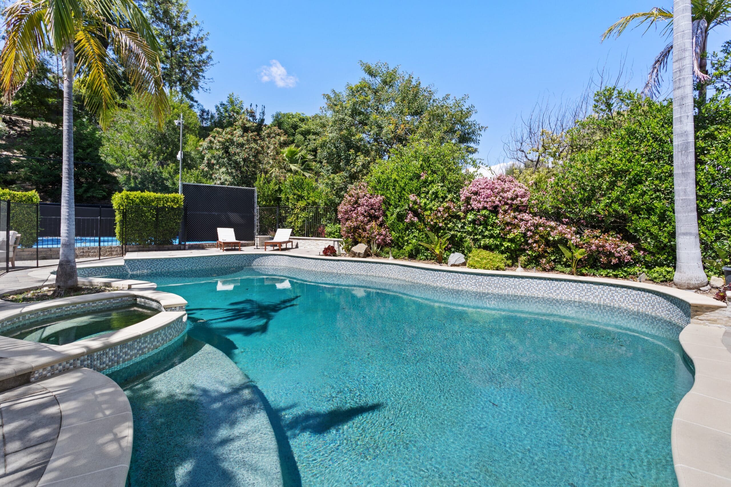 A clear blue swimming pool with an attached hot tub, surrounded by lush green trees and blooming bushes, with lounge chairs and a tennis court visible in the background under a sunny sky.