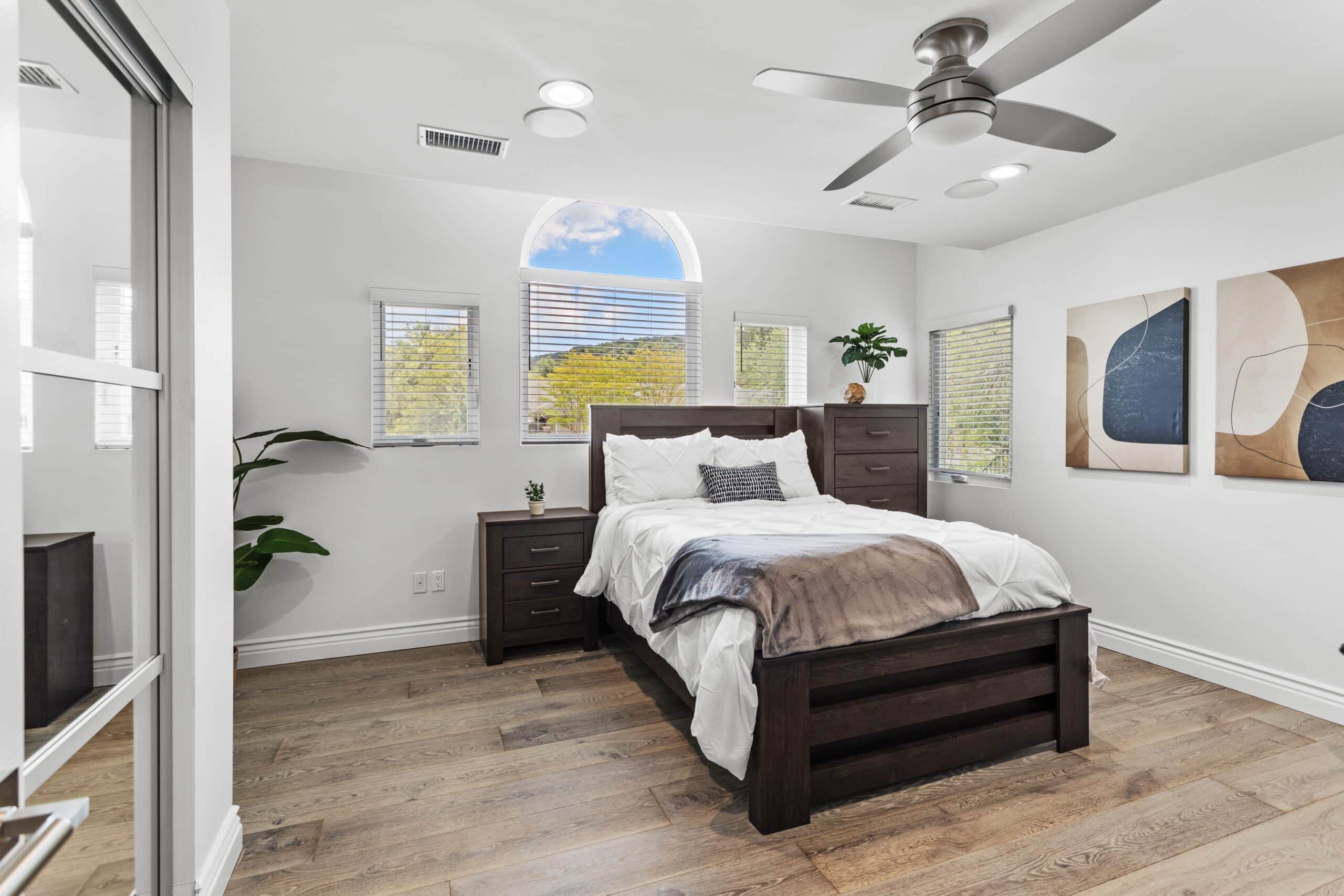 Modern bedroom with a wooden bed frame, white bedding, nightstands, abstract wall art, wood floor, ceiling fan, potted plants, and windows letting in natural light with a view of trees and blue sky.