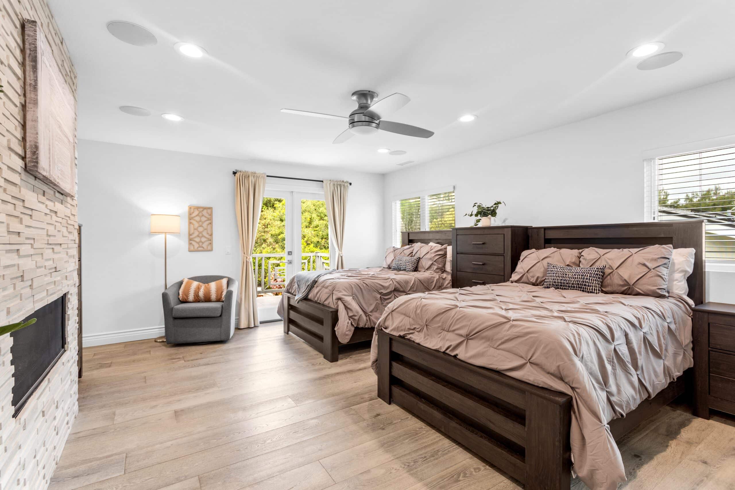 Spacious, bright bedroom with two dark wooden beds, beige bedding, a gray armchair, wood floors, a ceiling fan, large windows, and a sliding door opening to a balcony with greenery outside.