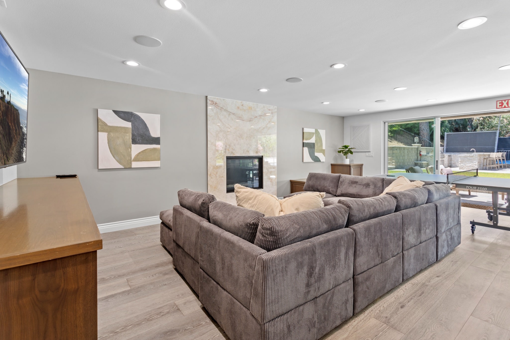 A modern living room with a large gray sectional sofa, abstract wall art, a marble fireplace, wood furniture, and sliding glass doors leading to a green backyard with outdoor seating.